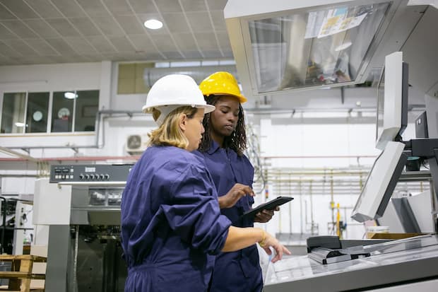 Female industrial employee teaching colleague to operate machine, pointing at control board, using tablet