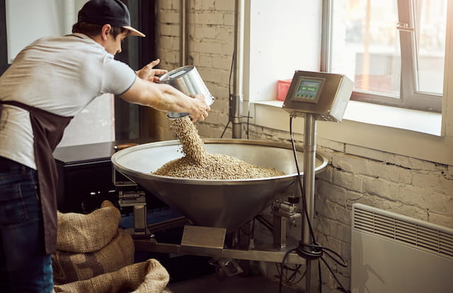 Young man in cap pouring green coffee beans into metal hopper while using electronic scales